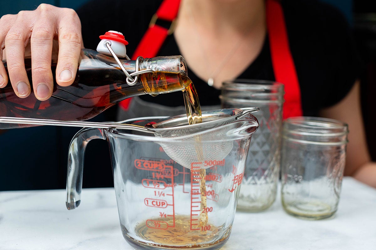 A baker pouring aged vanilla extract through a sieve into a measuring cup to filter out the vanilla bean.
