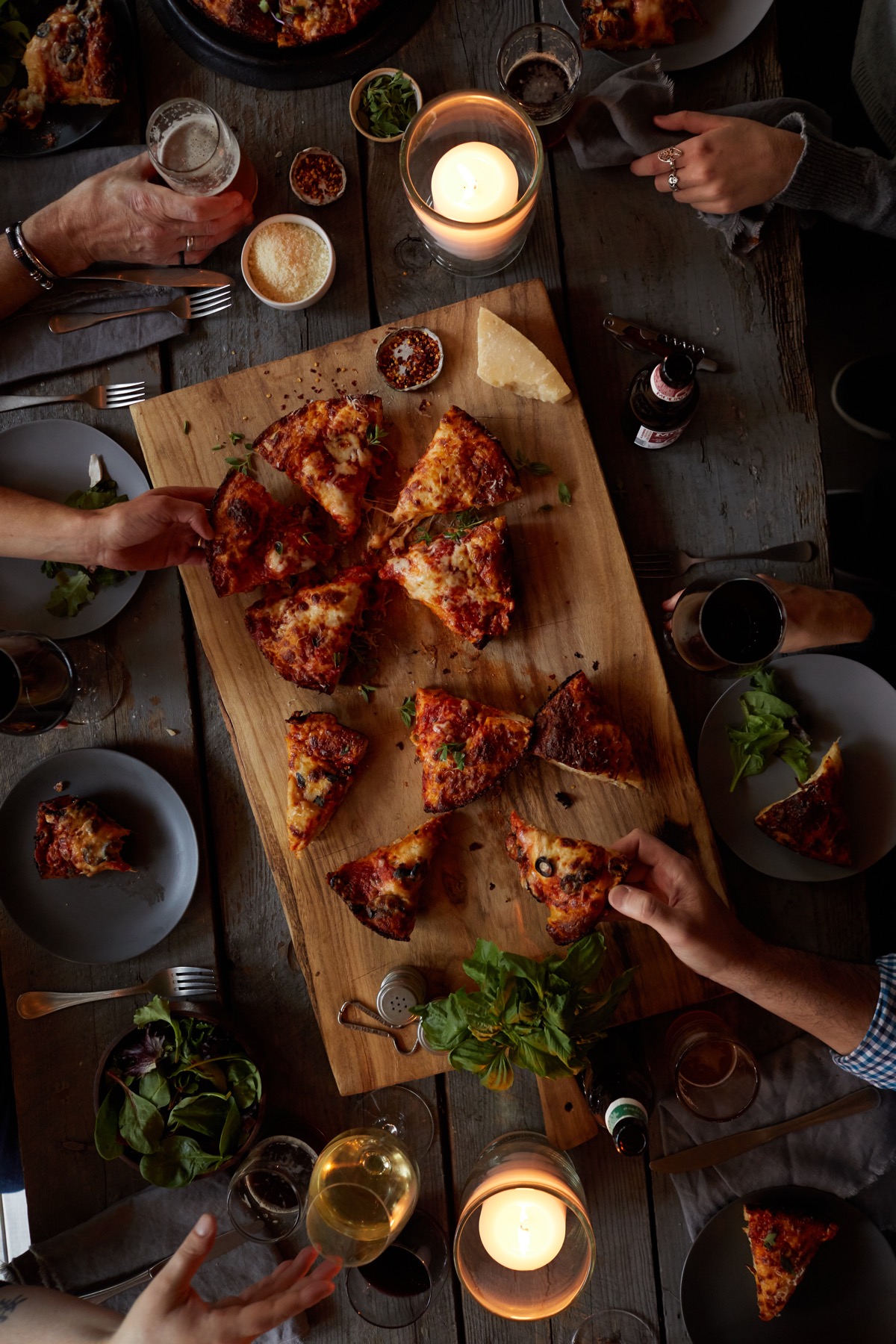 Baked pizza on a cutting board with lit candles, wine, beer, and hands of people sharing.