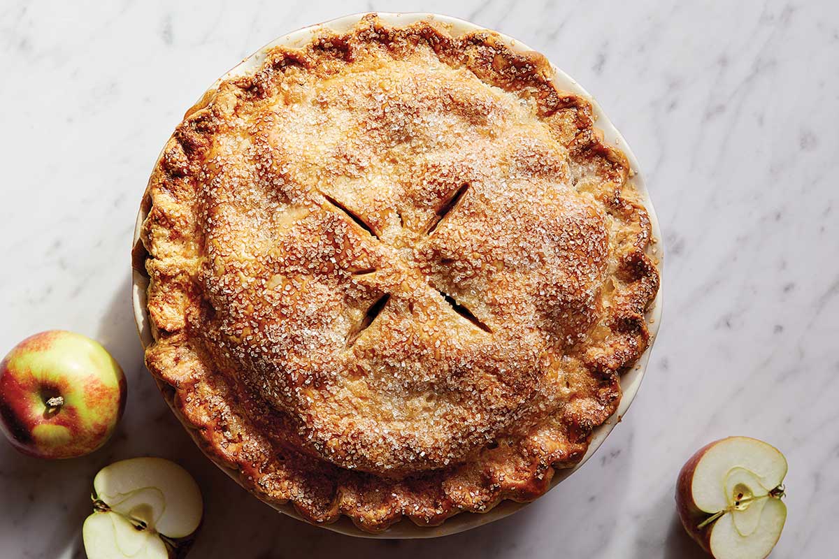 A traditional double crust apple pie topped with sparkling sugar next to a few apples