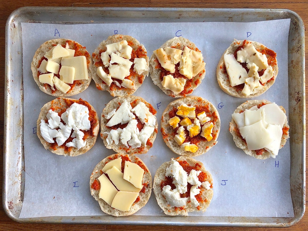 10 English muffin halves topped with tomato sauce, each one with a different type of cheese, ready to bake.