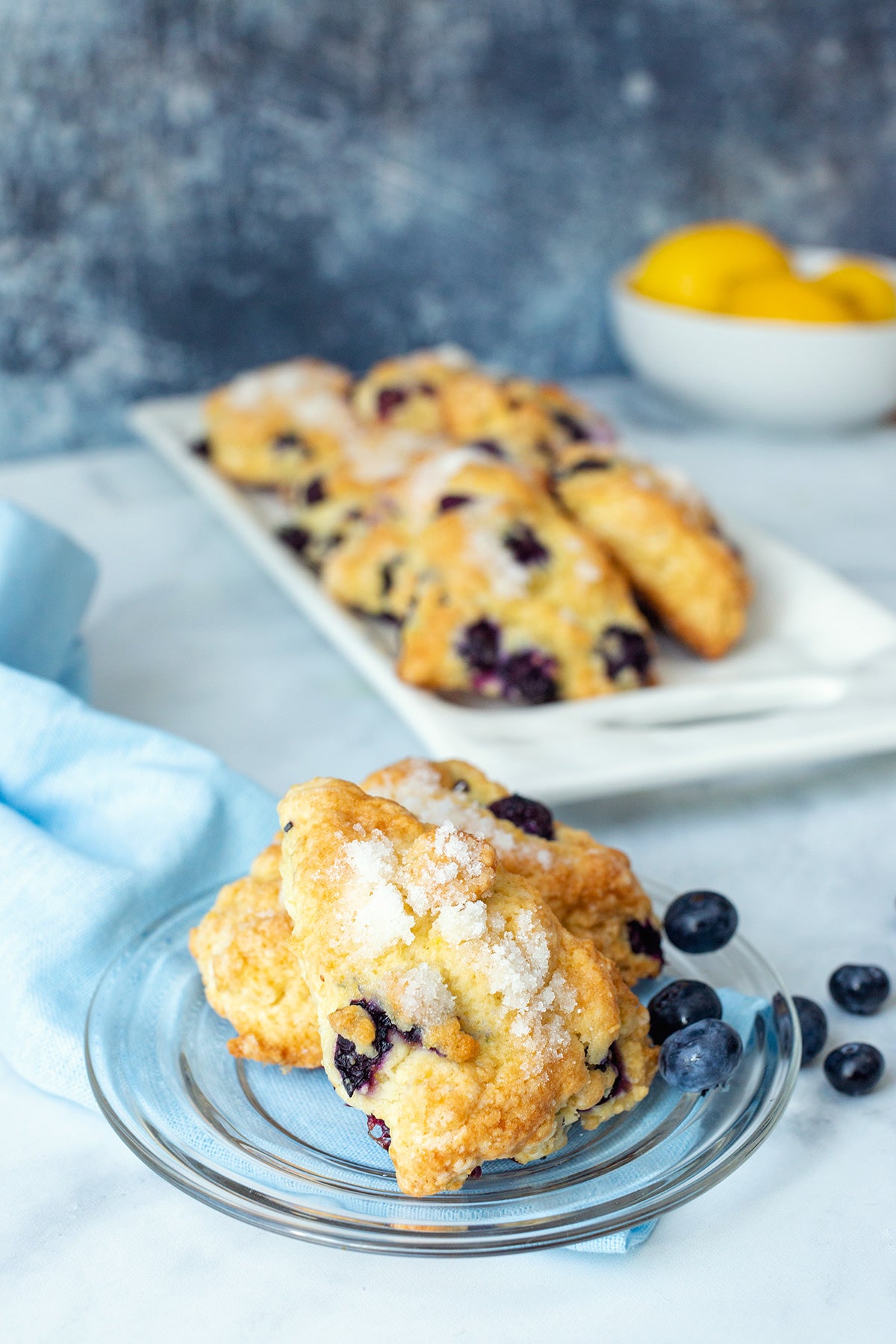 Fresh blueberry scones on a platter next to a bowl of lemons with two scones on a plate, ready for serving