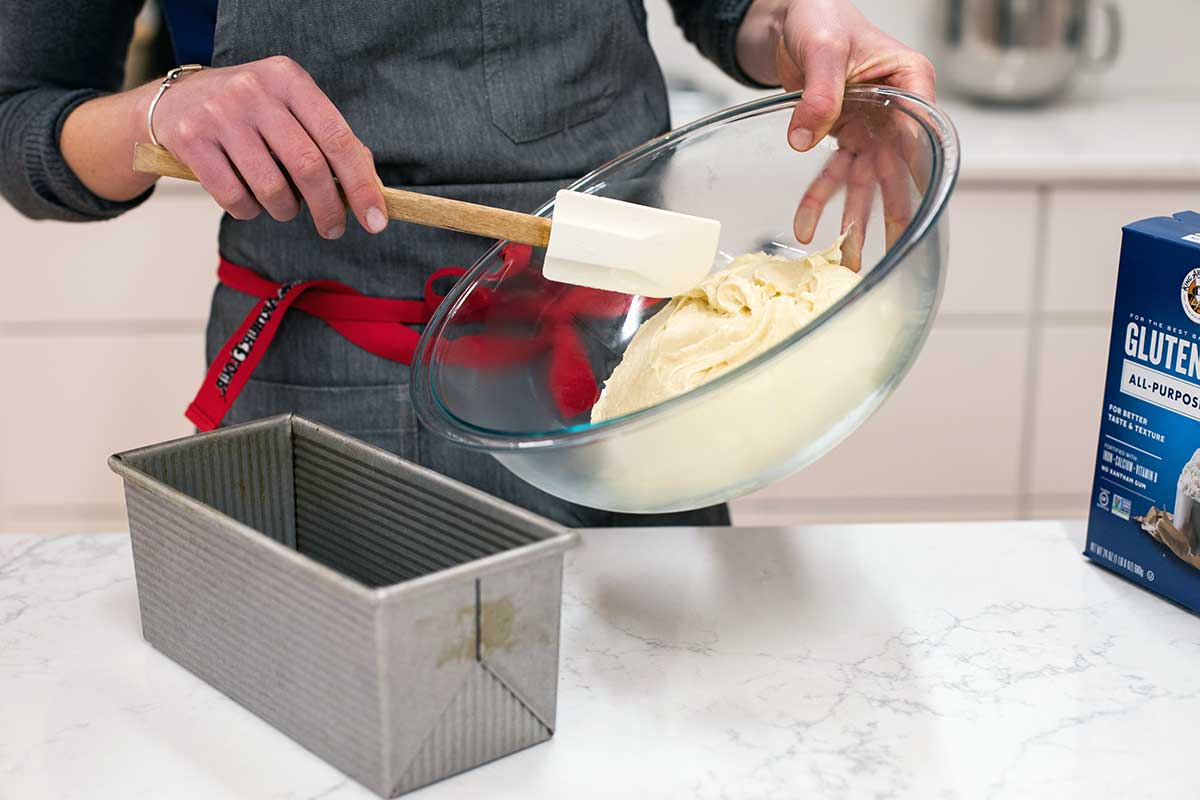 A baker pouring gluten-free bread dough from a bowl into a loaf pan for its second rise