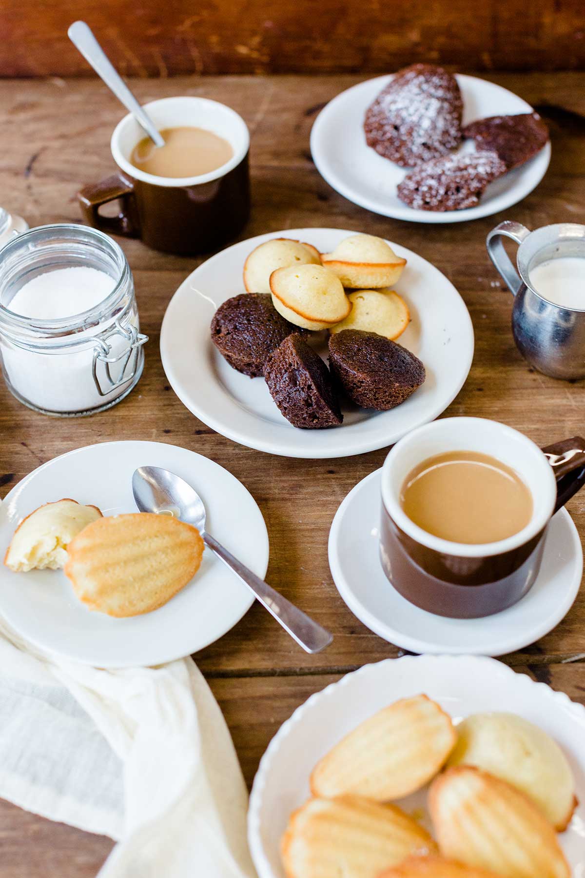 A table arranged with a platter of madeleines, coffee cups, plates, spoons, and a jar of sugar