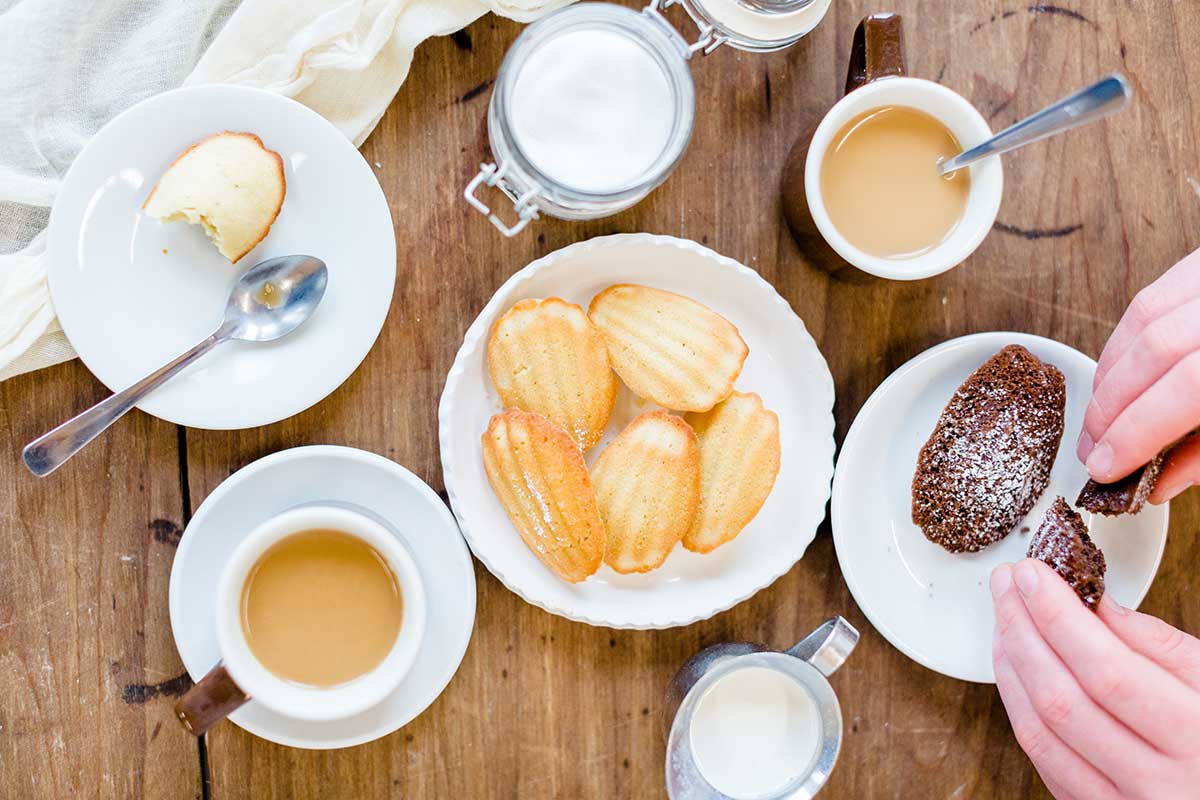 A plate of vanilla madeleines on a wooden table with a few cups of coffee, tea, and small plates