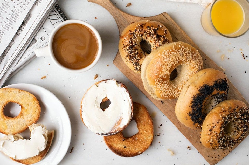 A bread board with homemade bagels next to a cup of coffee, orange juice, and a toasted bagel with cream cheese