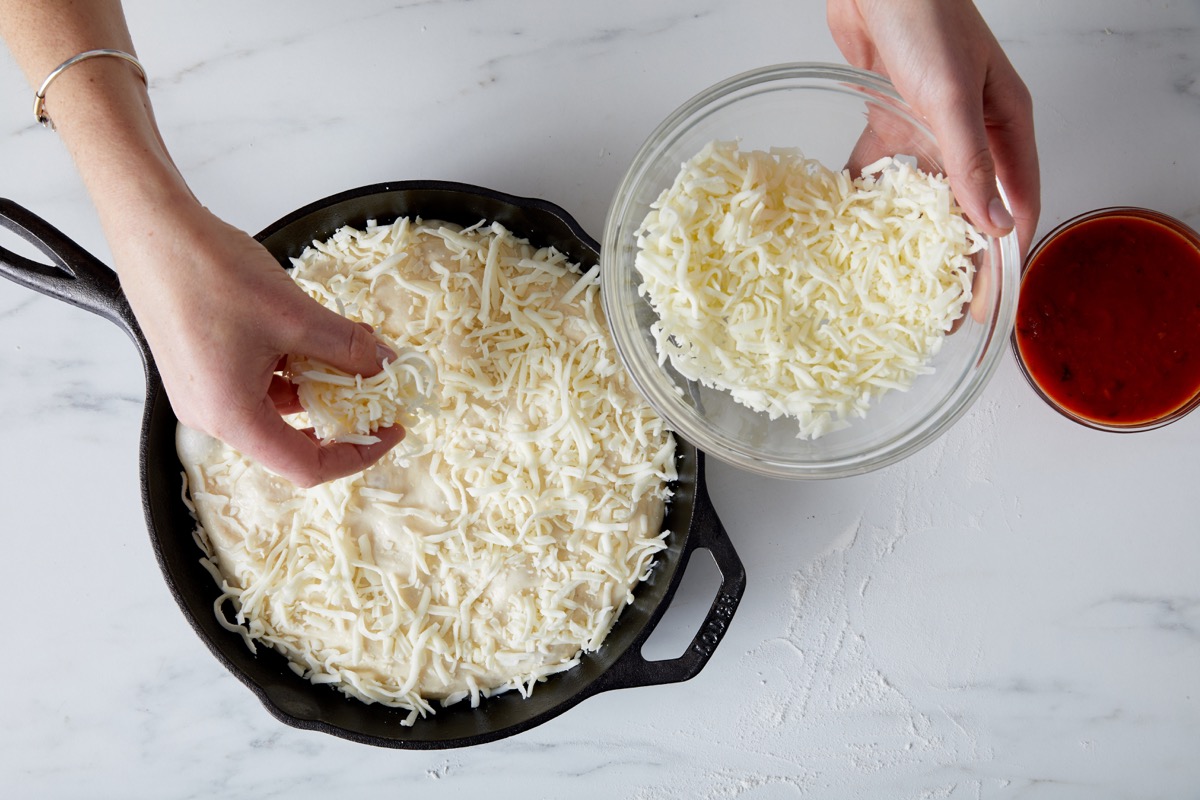 Cheese being sprinkled on unbaked pizza crust in a cast iron pan.