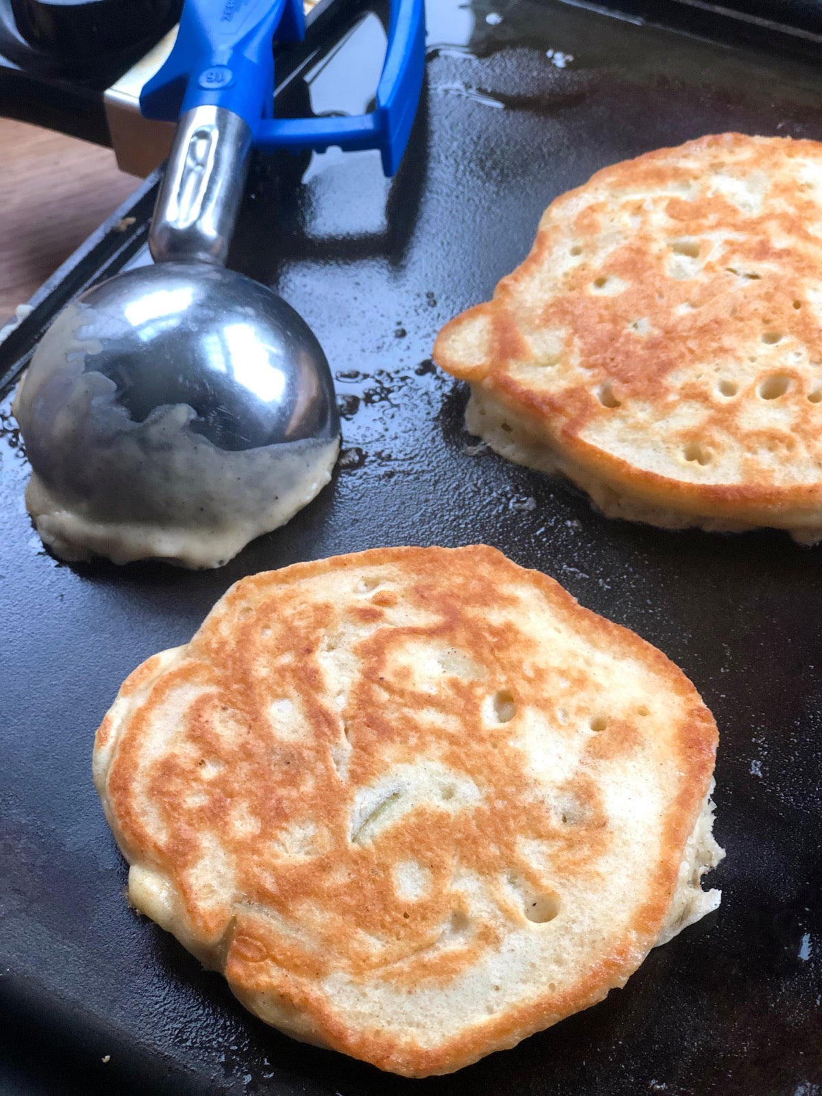 Two pancakes cooking on a griddle, muffin scoop on the side.