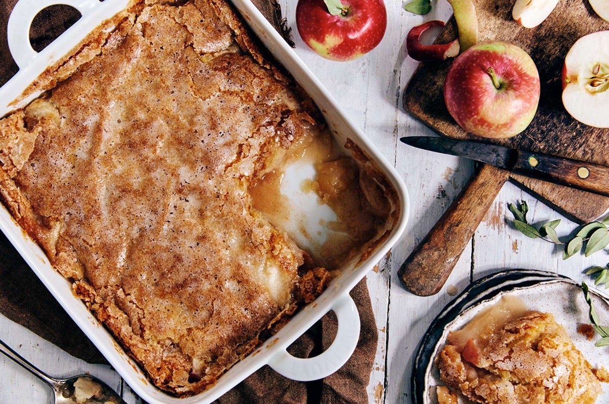 Sugar-crusted apple cobbler in a pan, some on a plate, apples on the table alongside.