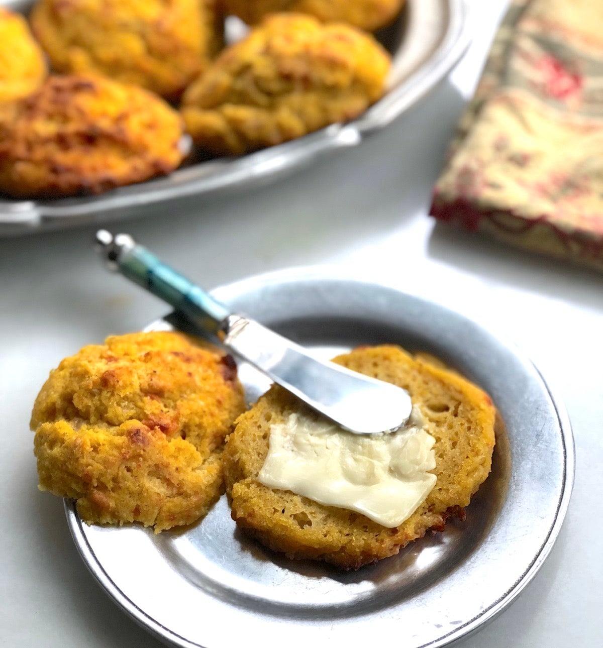 Pumpkin cheddar biscuit on a plate, spread with soft butter, more biscuits in the background.
