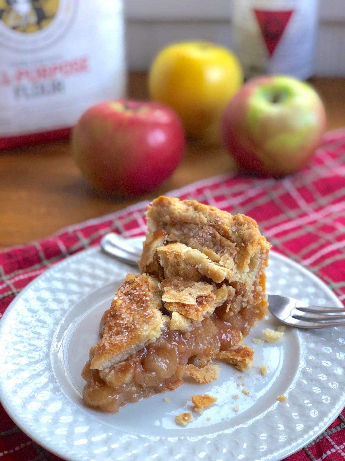 Slice of apple pie on a plate, apples in the background.