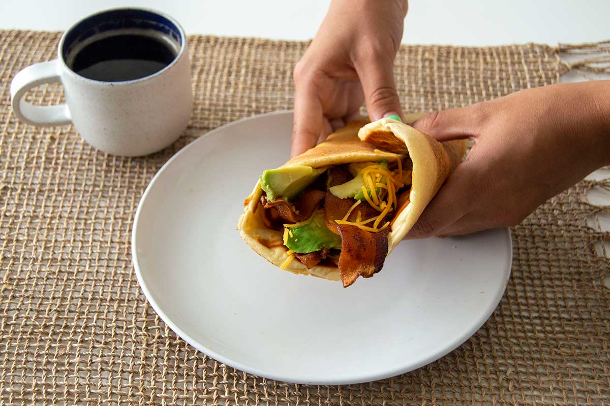 Hands holding bacon and avocado wrap, with a pancake as the flatbread