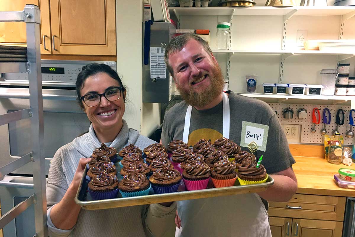 Two bakers holding up a tray of frosted cupcakes, smiling
