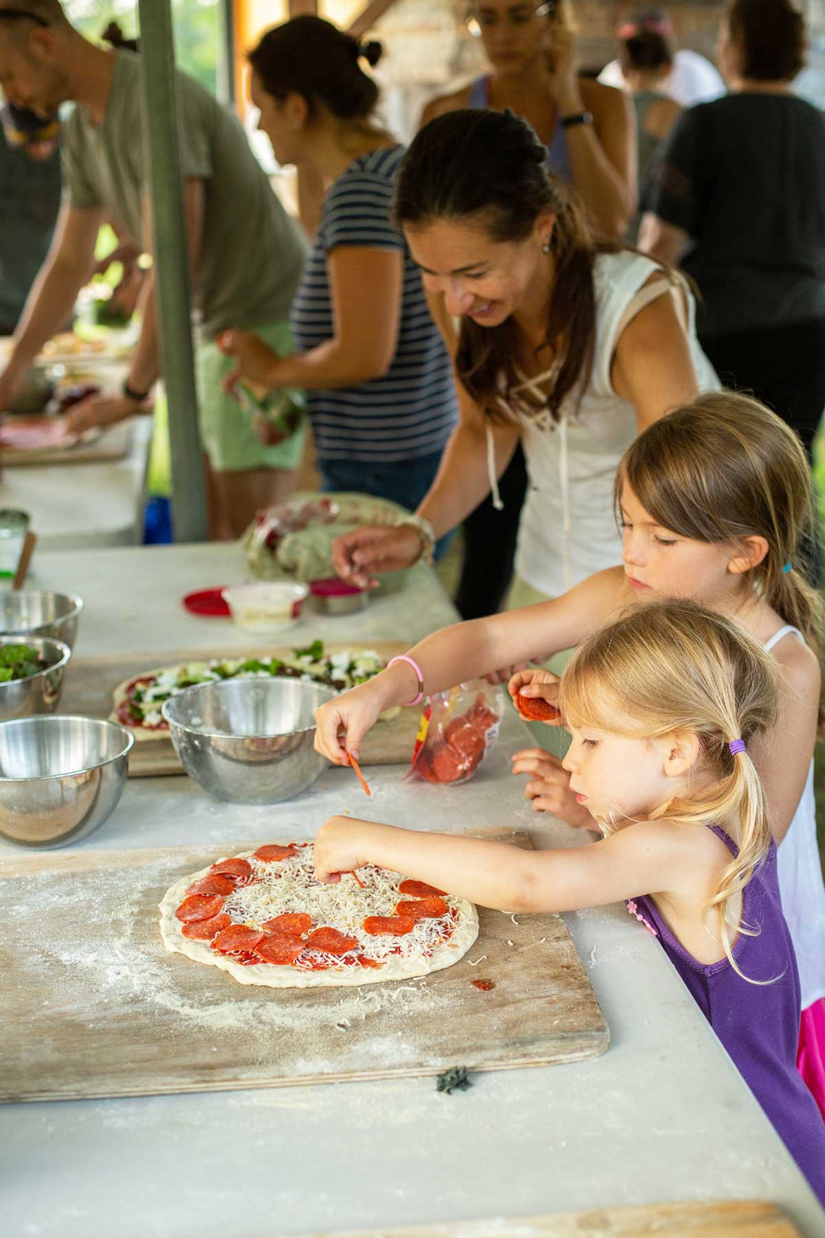 Two small children helping top a pizza before it goes into an outdoor oven