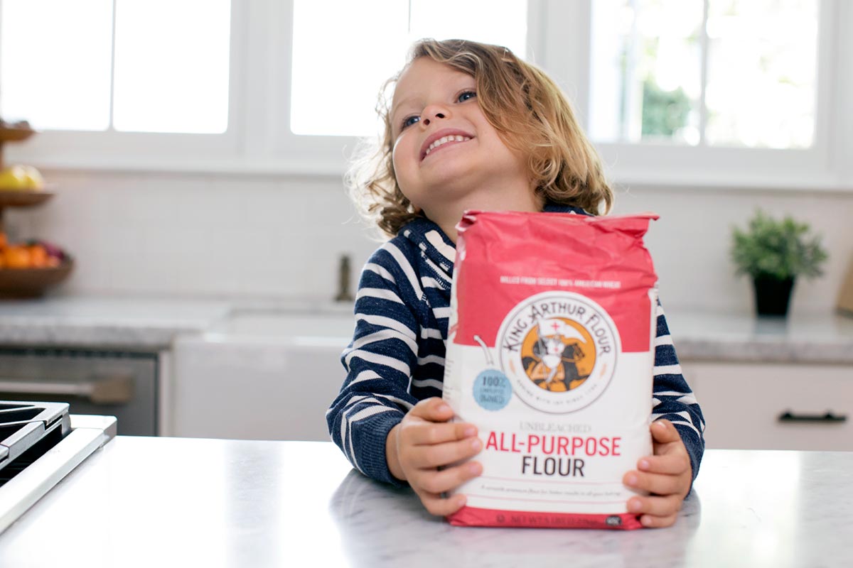 A small child smiling with a bag of flour in a kitchen