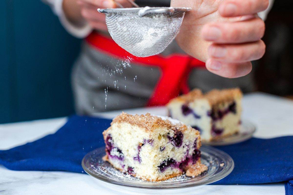 A baker dusting a slice of Blueberry Buckle Coffeecake with confectioners' sugar