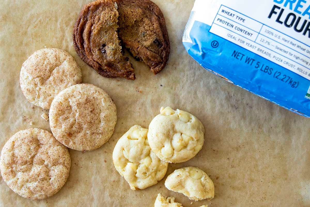 Buttery snickerdoodles, chocolate chip cookies, and white chocolate cookie drops on parchment next to a bag of bread flour