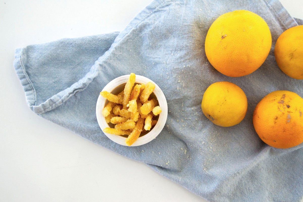 Small bowl of candied citrus peels next to three oranges