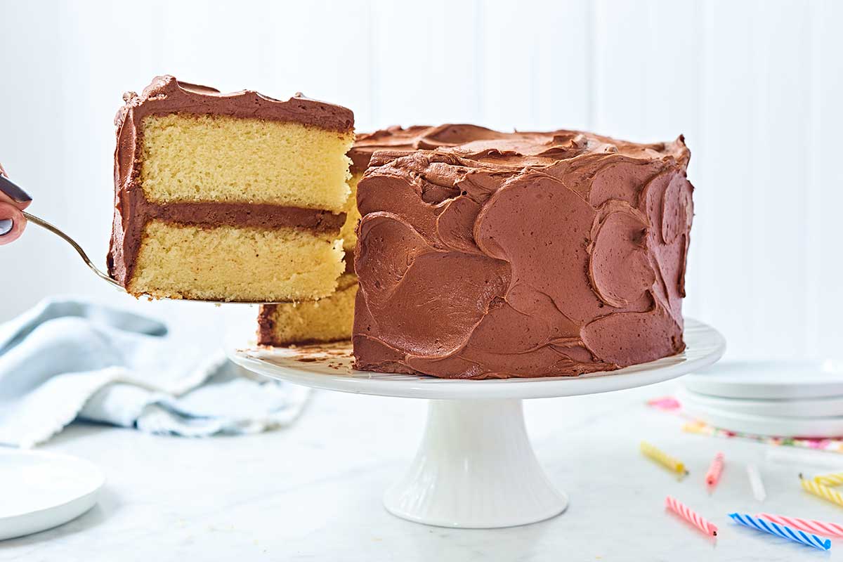 A slice being remove from a Classic Birthday Cake: yellow layer cake coated in chocolate fudge frosting
