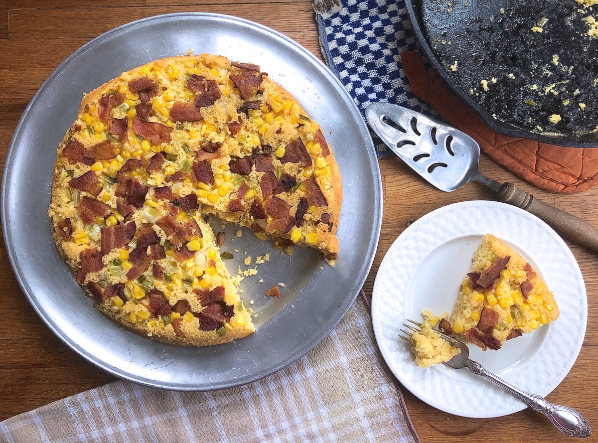 Baked cornbread in a skillet topped with bacon, scallions, and corn; one wedge on a serving plate