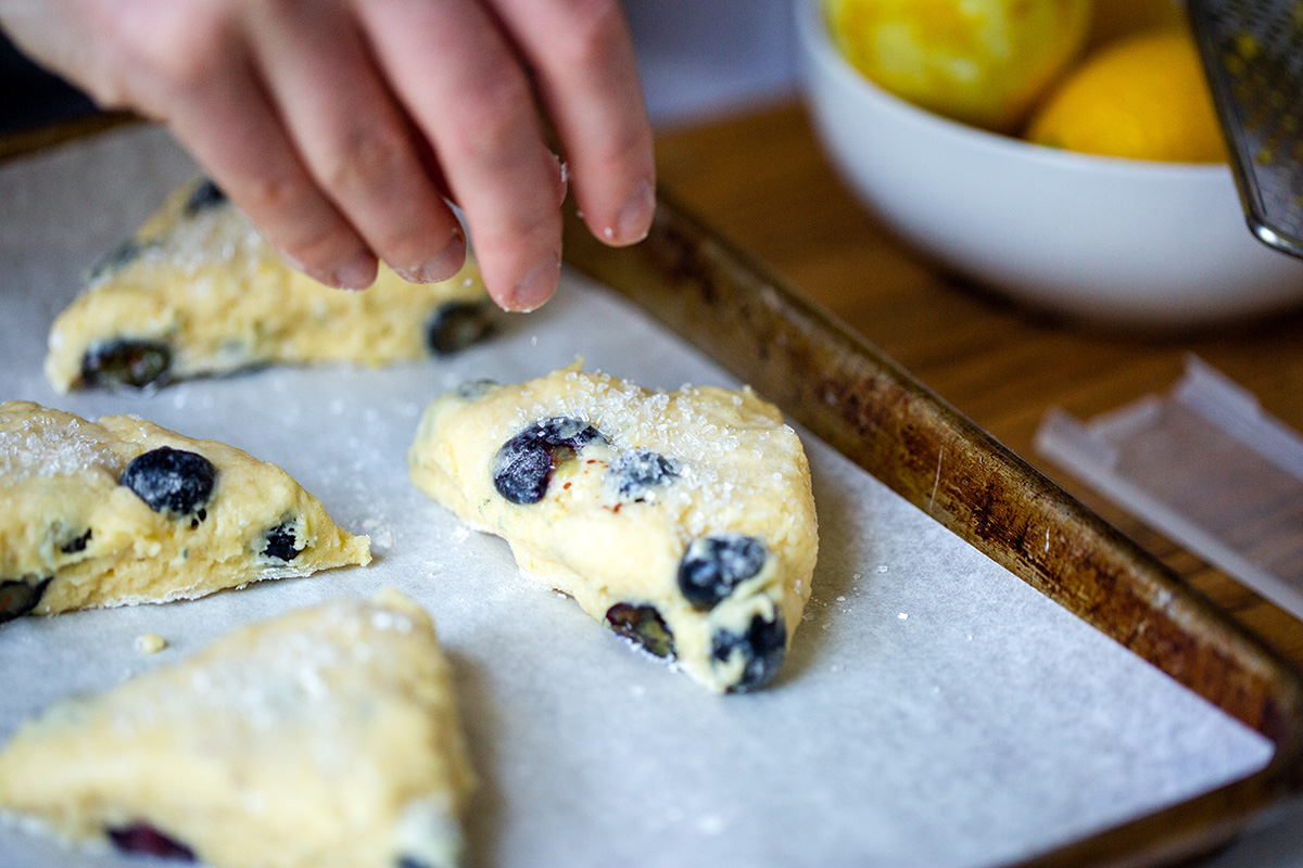 A baker's hand sprinkling unbaked blueberry scones with sparkling sugar on a baking sheet