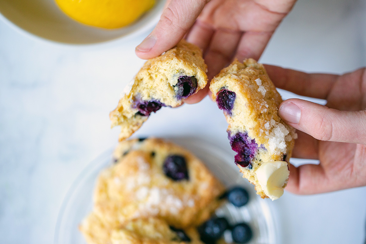 A baker's hand holding out a fresh blueberry scone, broken in half, showing the texture with a few lemons in the background