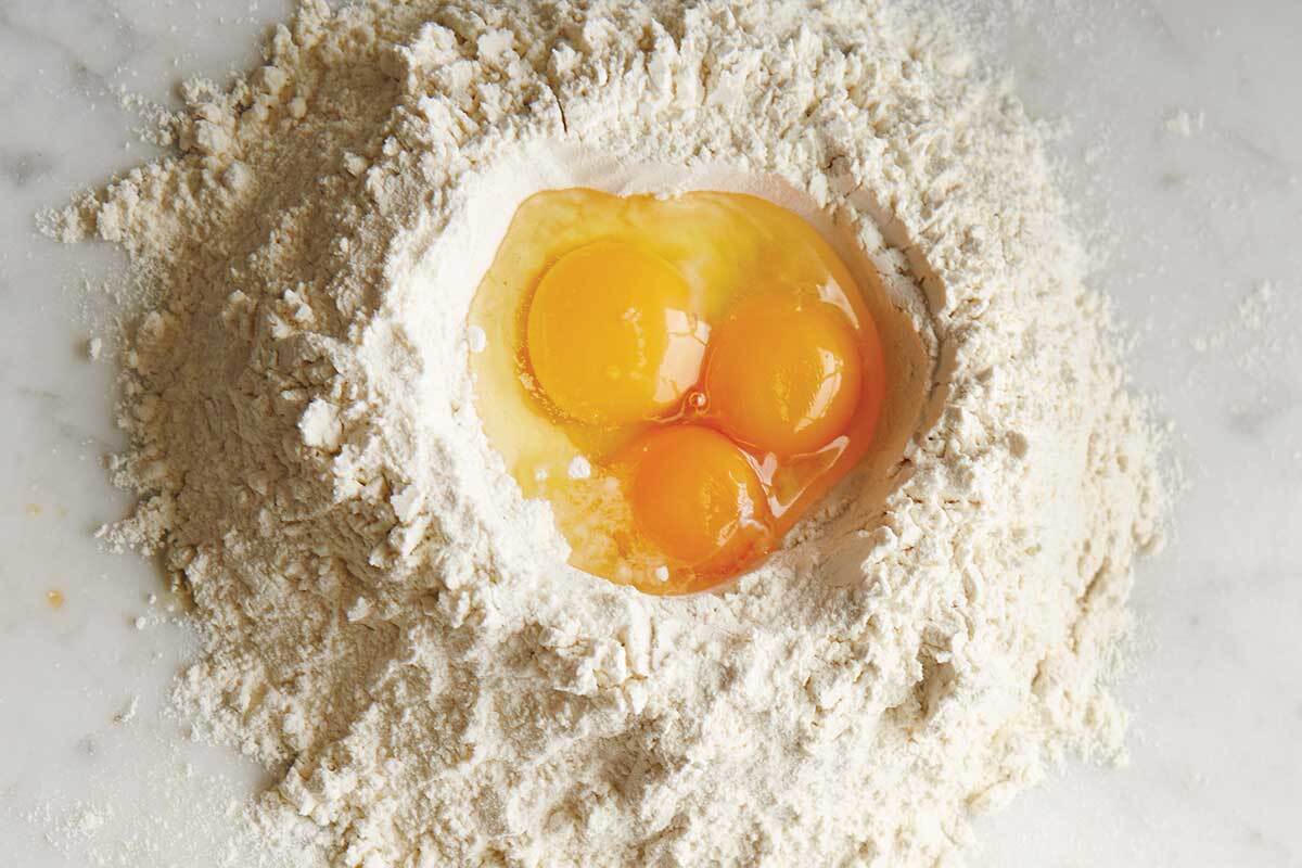 Three eggs cracked into the middle of a pile of pasta flour, about to be mixed into dough