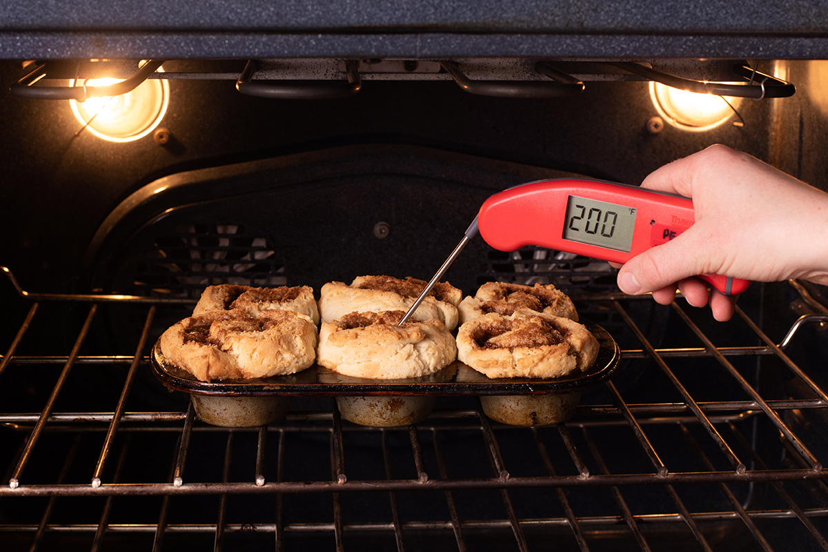 A baker testing the temperatuer of gluten-free cinnamon rolls baking in the oven, showing that it's about 200 degrees F
