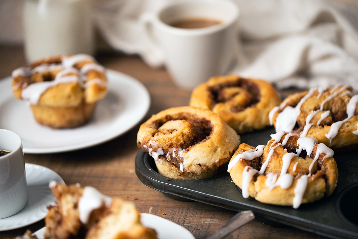 Gluten-free cinnamon rolls on a tray next to a cup of coffee and plate with a cinnamon roll, ready to be enjoyed