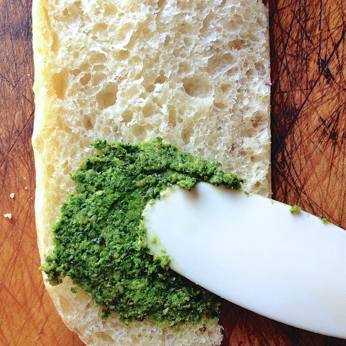 Bright green pesto being spread on a slice of baguette.