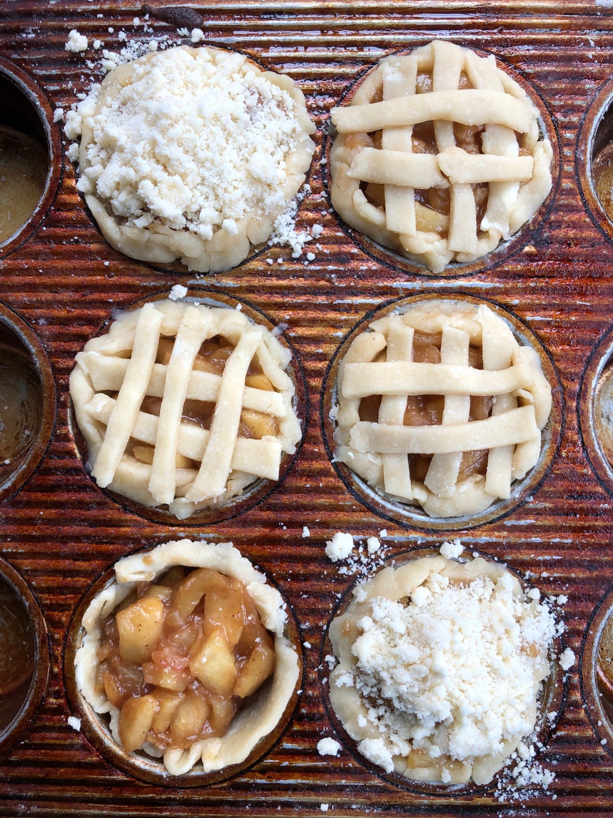 Six mini pies in a muffin pan, ready to bake, showing lattice top crusts and crumb top crusts.