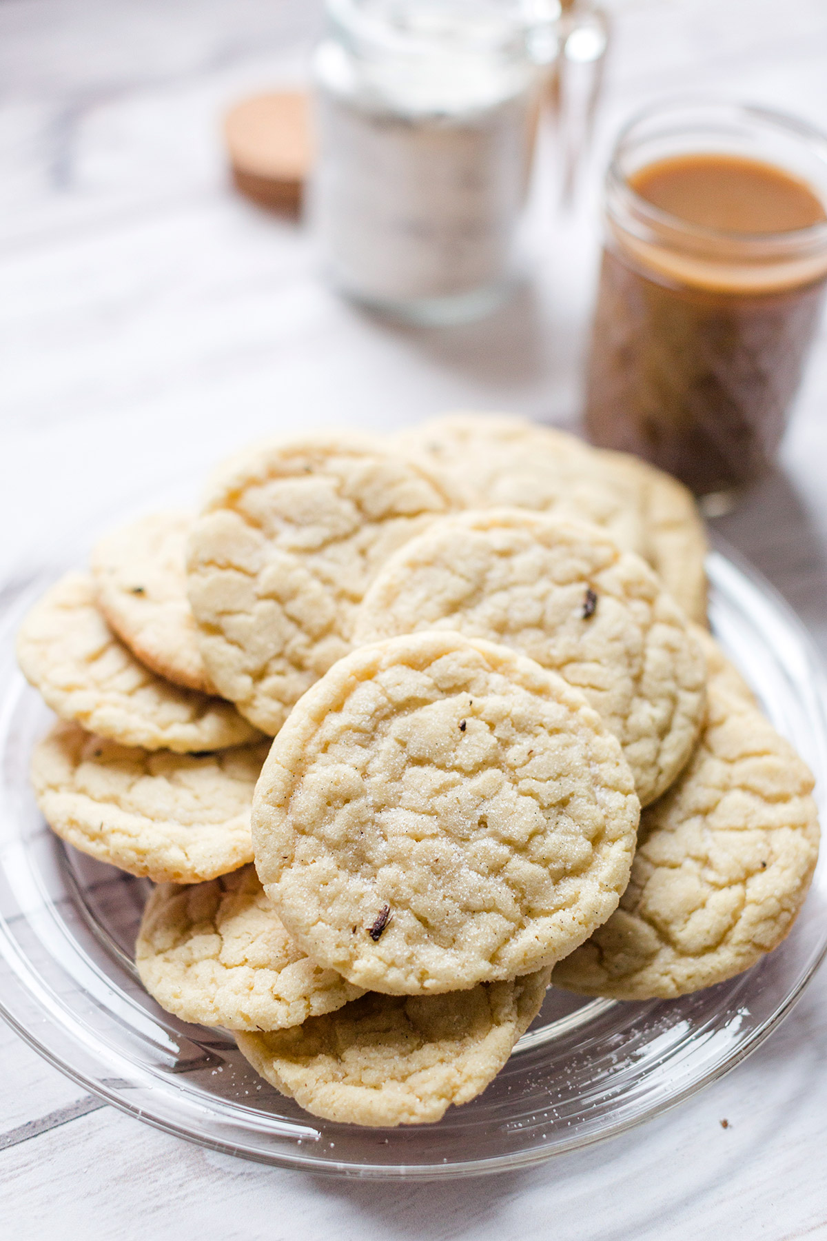 A plate of vanilla bean sugar cookies next to a cup of coffee and a jar of vanilla sugar