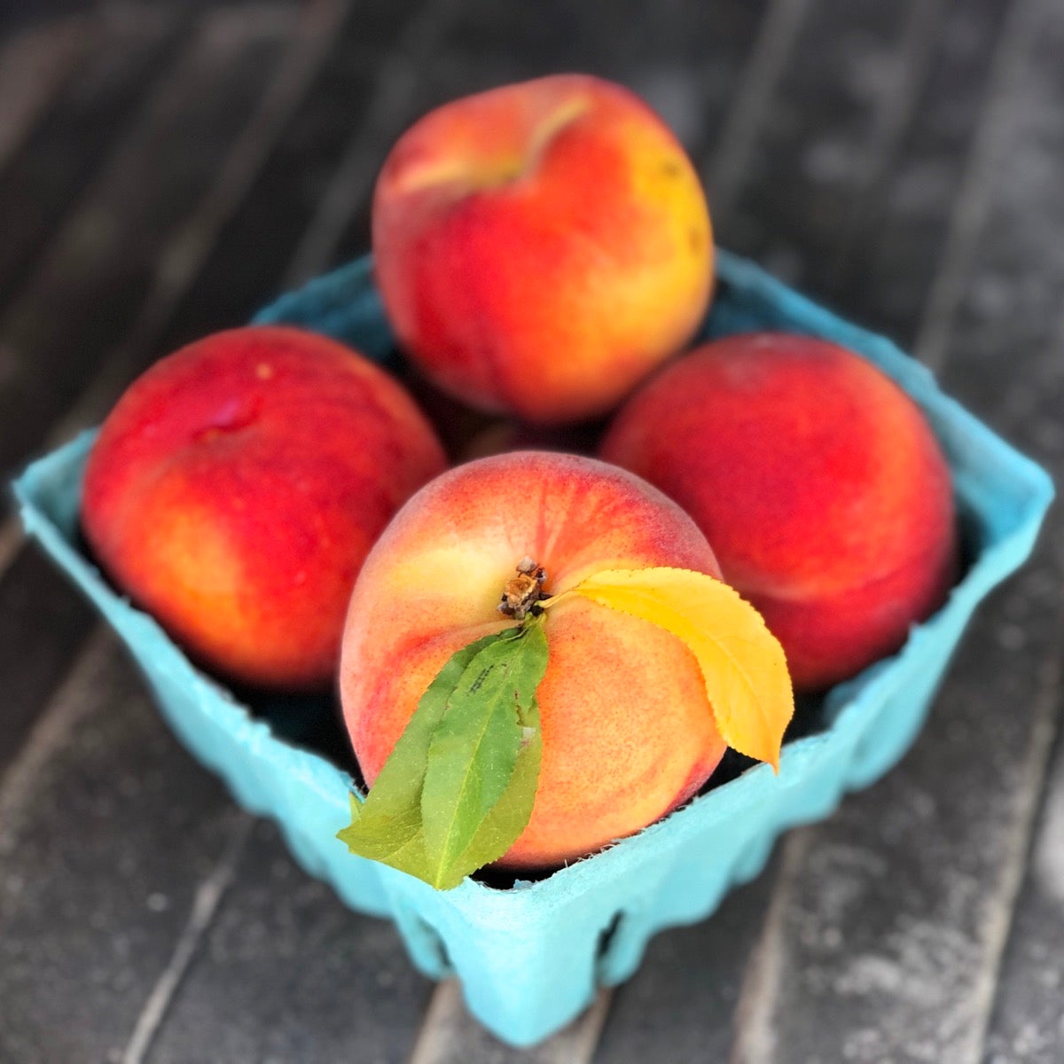 A quart container of beautiful fresh peaches, one with the leaves intact.