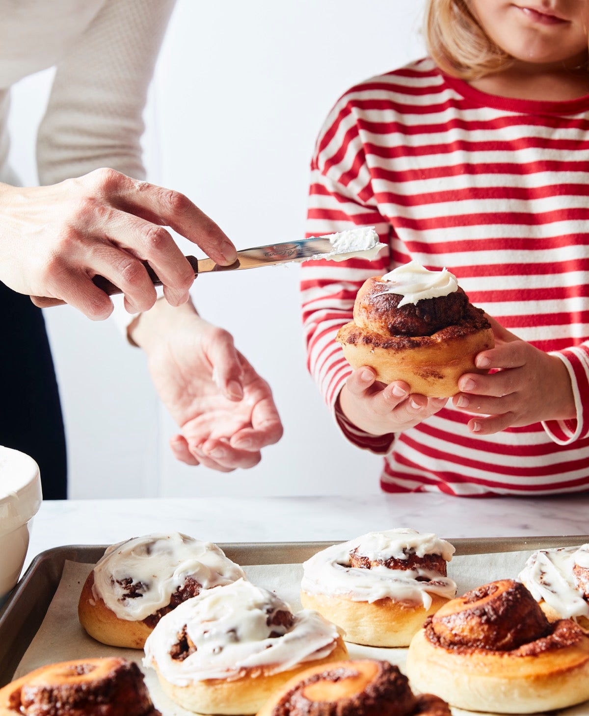 Little girl in a red-striped shirt holding a cinnamon roll while Mom frosts it with white icing.