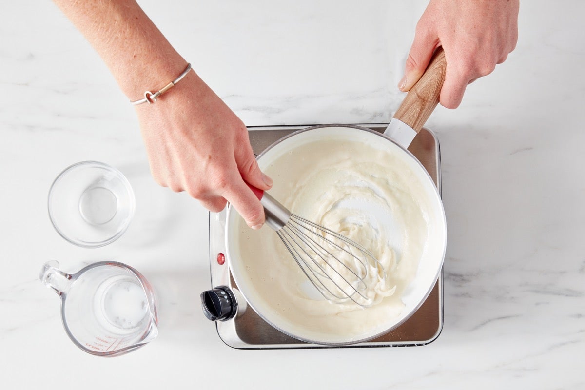 Hands using a whisk to mix flour and milk in a saucepan set over a portable burner.