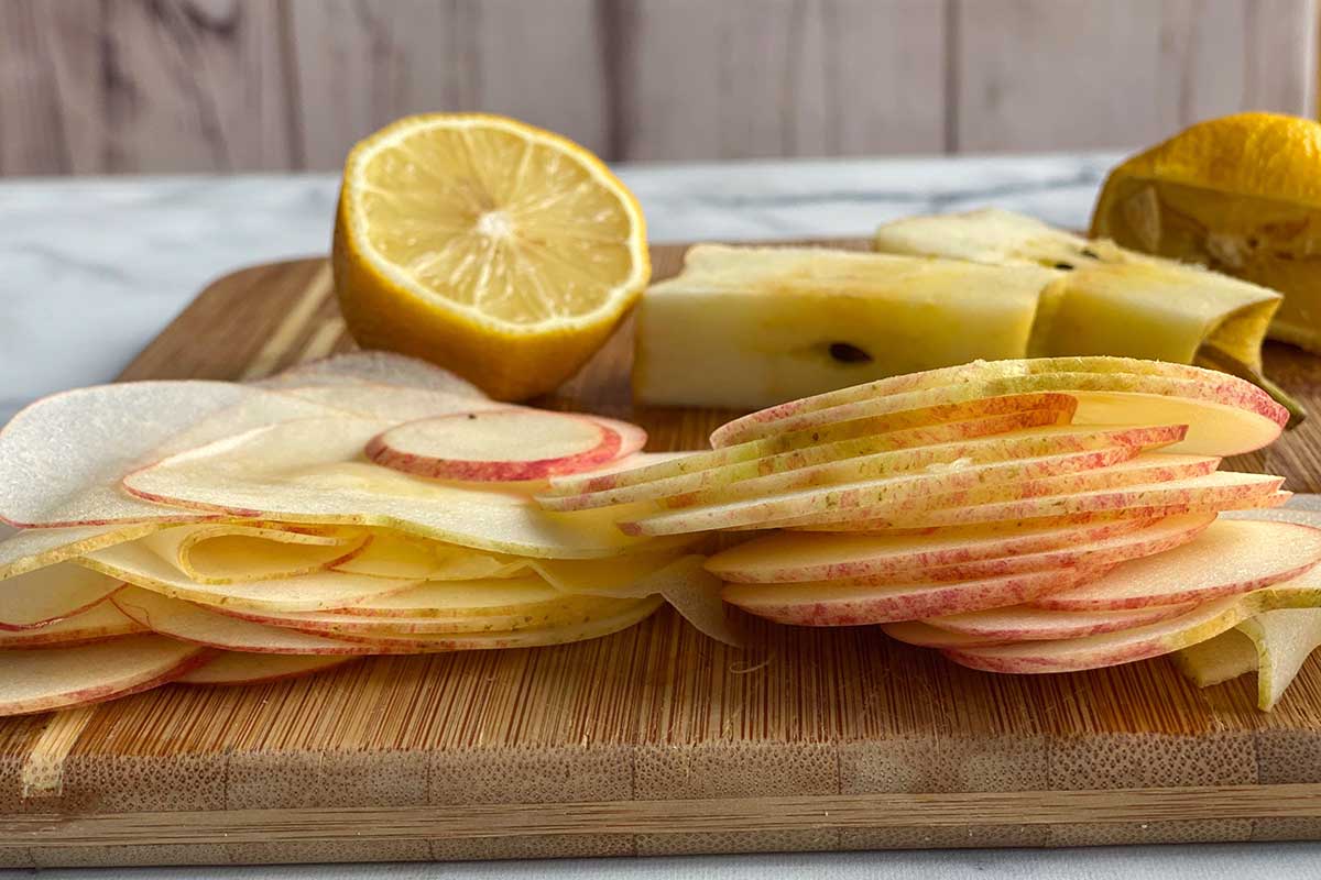A pile of thinly sliced apple discs on a wooden cutting board next to a lemon