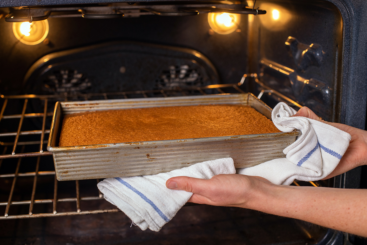 A baker pulling a vanilla sheet cake out of the oven to check for doneness