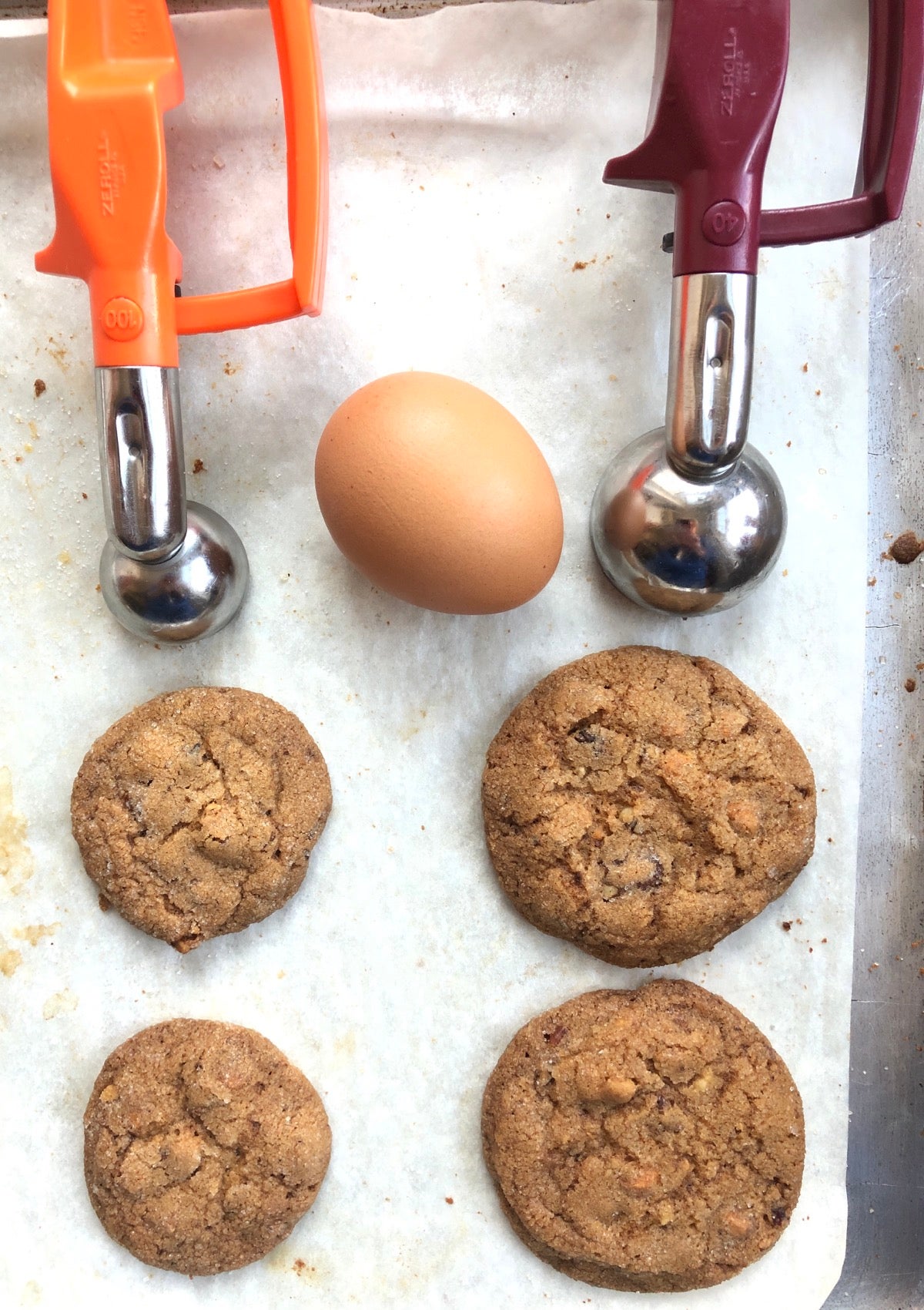 Two cookies on a baking sheet, one made with a tablespoon scoop, a smaller one made with a teaspoon scoop.