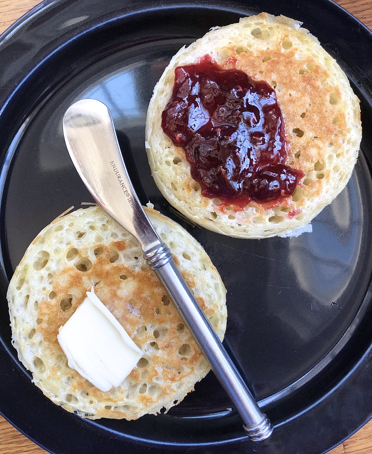 Two sourdough crumpets on a plate; one buttered, one spread with jam.