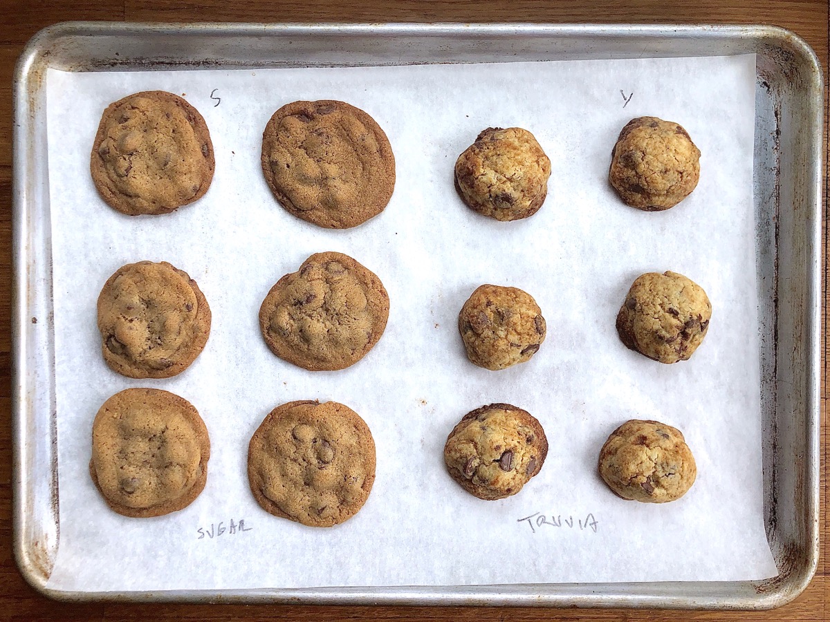 Chocolate chip cookies fresh from the oven on a baking sheet — half made with granulated sugar, half with Truvia, showing degrees of spread.