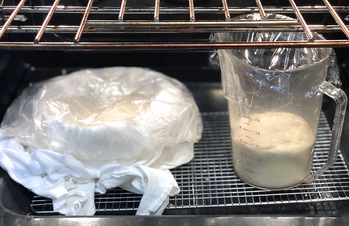 Bread dough rising in a large measuring cup; shaped loaf rising in a towel-lined bowl, both in a turned-off oven.