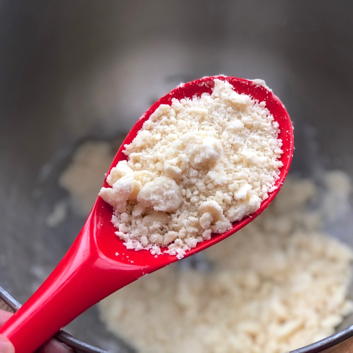 Butter and bacon fat worked into a bowl of dry ingredients to create a crumbly mixture, shown pictured in the bowl of a red spoon.
