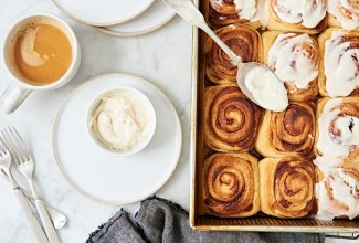 A tray of cinnamon buns with a bowl of frosting next to a cup of coffee