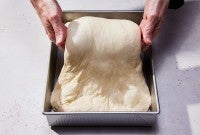 A baker folding wet dough in a square pan with their hands