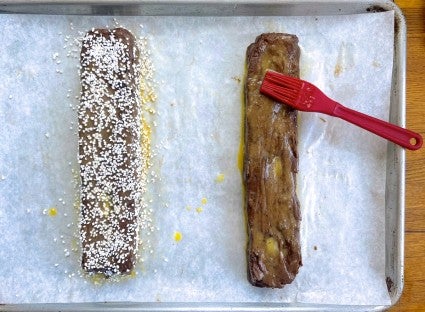 Malted Brownie Biscotti dough shaped into logs, being brushed with beaten egg and sprinkled with Swedish pearl sugar.