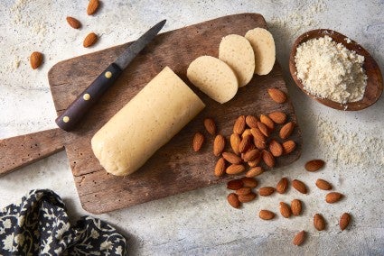 Homemade almond paste shaped into a log and sliced on a cutting board, surrounded by unskinned almonds.