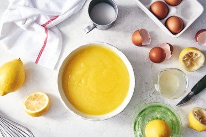 Lemon curd in a bowl surrounded by eggs, squeezed lemon halves, and a hand-held juicer.