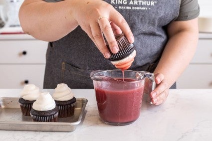 A baker dipping a frosted cupcake into a measuring cup of strawberry glaze