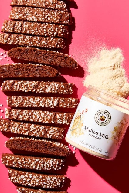 Chocolate malted milk biscotti, sliced and topped with sparkling sugar, next to a jar of malted milk powder