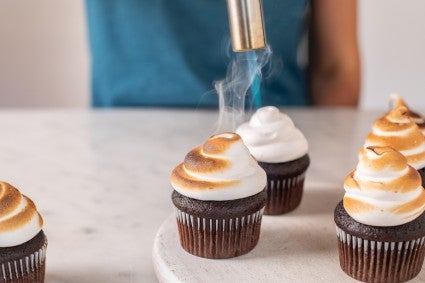 A baker using a torch to toast meringue-frosted cupcakes