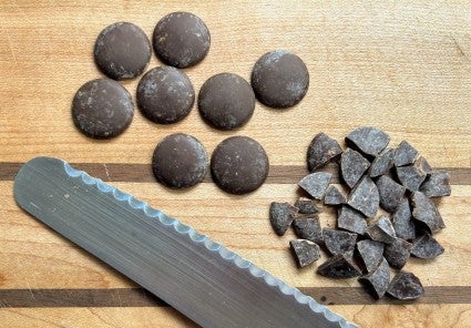 Whole chocolate disks and disks chopped into chopped into quarters on a wooden cutting board, with end of serrated knife showing.